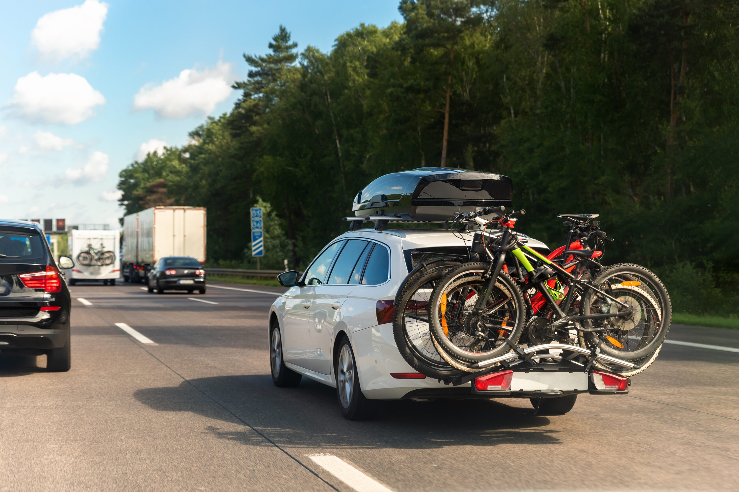 travel bike rack car Back view modern white familywagon car with mounted roof box trunk and bike tail carrier driving european highway road against blue sky summer day. Lifestyle travel adventure trip journey concept.