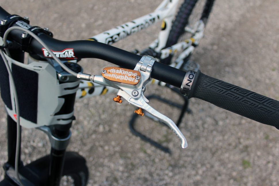 Close-up view of a mountain bike handlebar showing a brake lever with 'making up the numbers' engraved on it, along with the grip and part of the bike frame in the background.