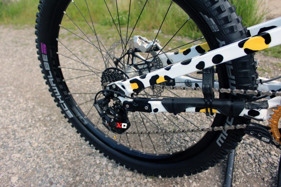 Close-up view of a mountain bike's rear drive train, showcasing a unique black and yellow polka dot design on the frame. The bike is positioned on a gravel surface with greenery in the background.