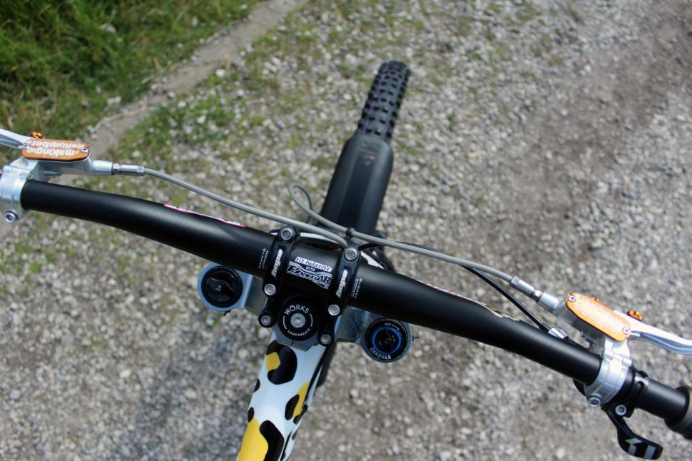 Top view of a mountain bike's handlebars with a black stem and yellow and black patterned frame, showcasing the brake and shifter levers.
