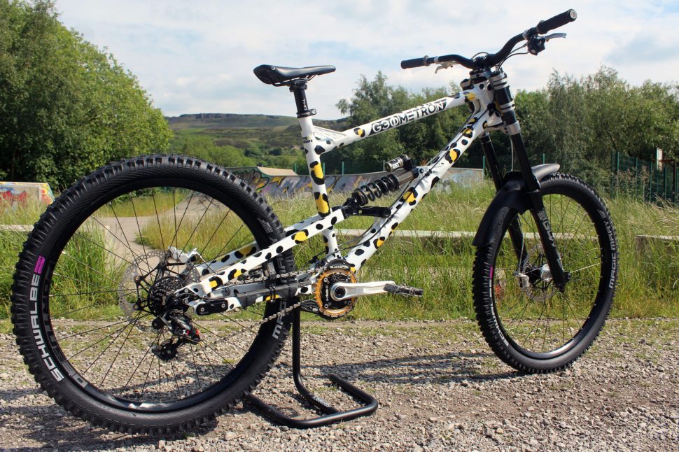 A Geometron mountain bike with a distinctive polka dot design, resting on a stand in a grassy area, with a backdrop of a trail and trees.