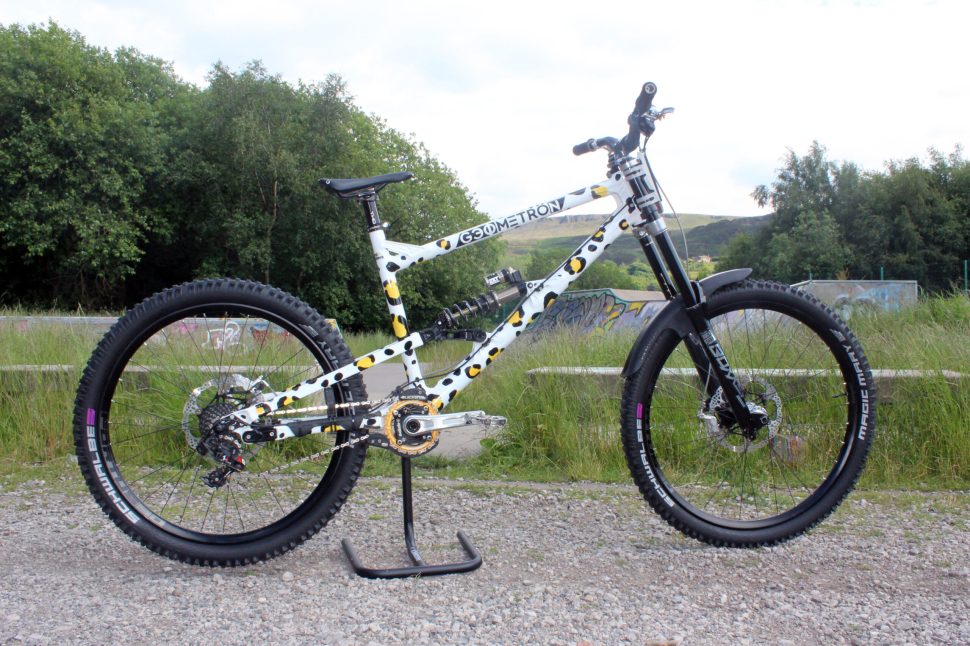 A side view of a Geometron mountain bike featuring a unique black and white speckled design, positioned on a bike stand in a grassy area with trees in the background.
