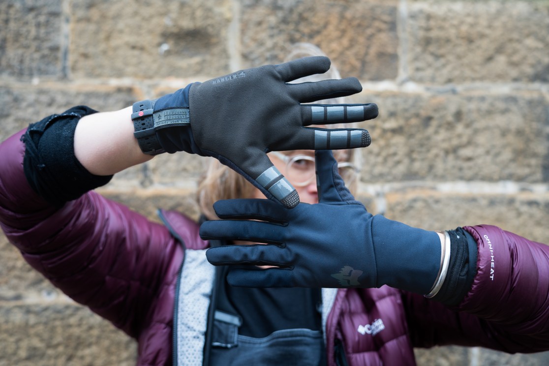 A person showing two different types of gloves, one on each hand, against a stone wall background. The left hand displays a glove with reflective elements, while the right hand shows a solid-colored glove.