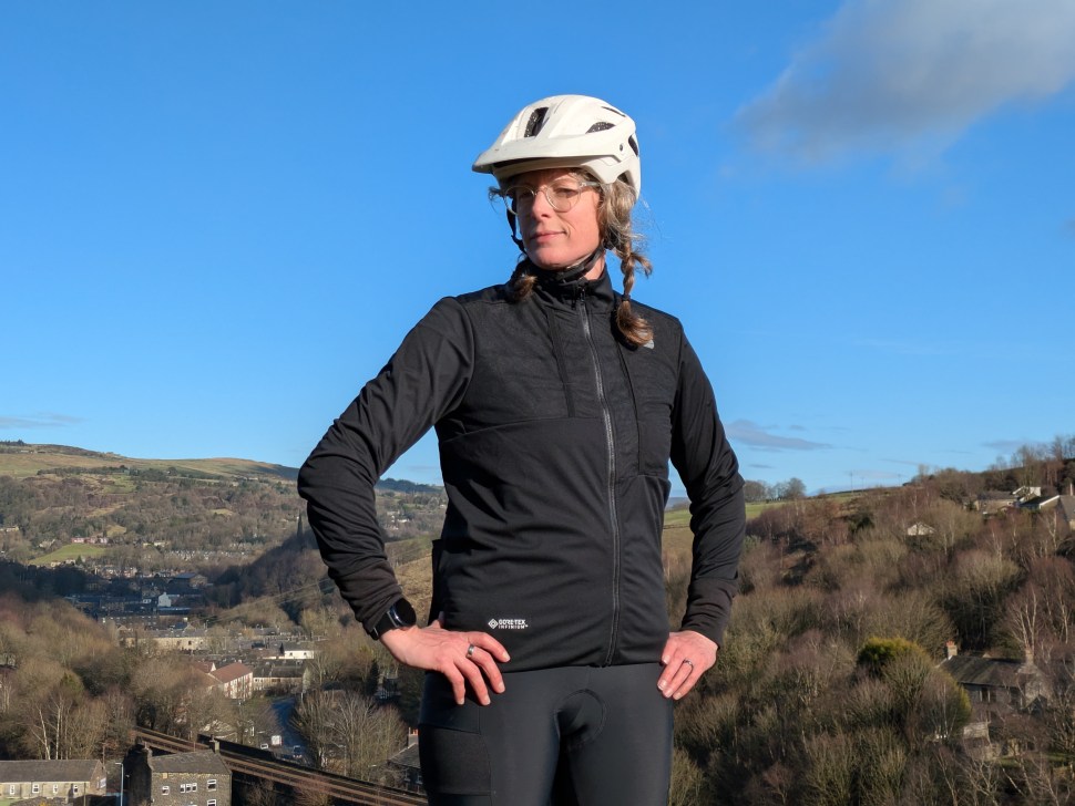 A cyclist stands outdoors wearing a black cycling jacket and a white helmet, with a scenic hilly landscape in the background.