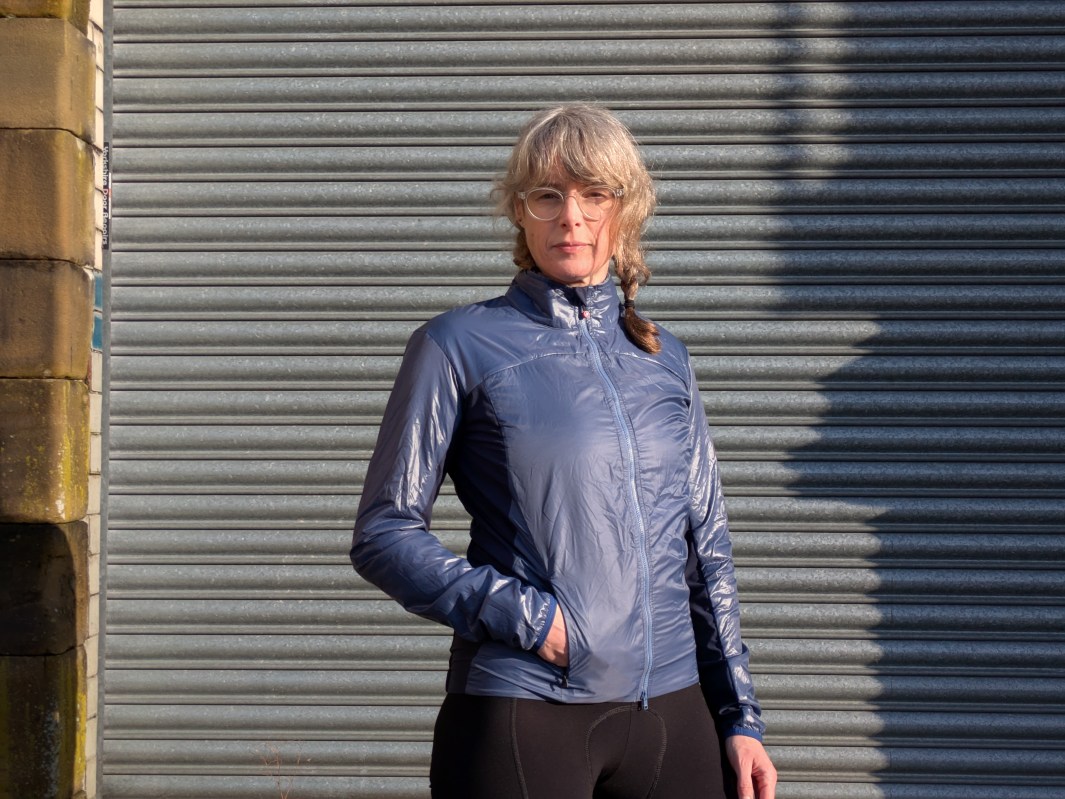A person wearing a shiny blue jacket stands confidently in front of a grey corrugated background, with one hand in the jacket pocket.