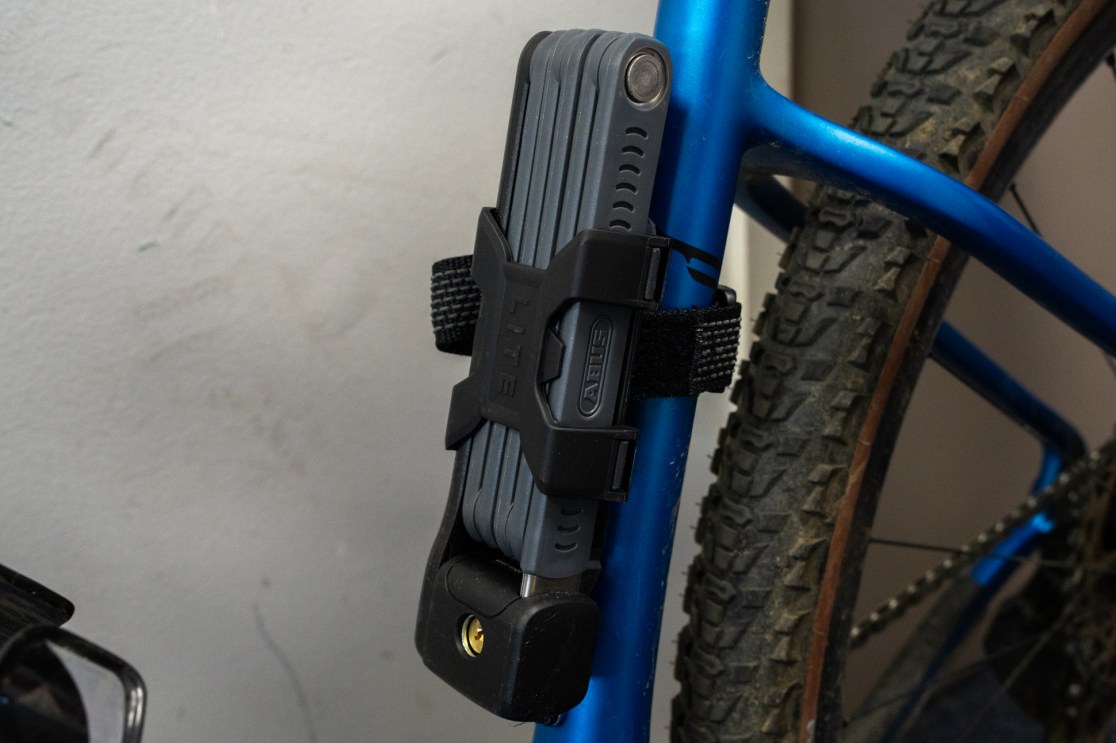 Close-up image of a bike frame with an Abus folding lock securely attached using a black strap. The bike has a blue metallic finish and features knobby tires, suggesting it is designed for off-road use.
