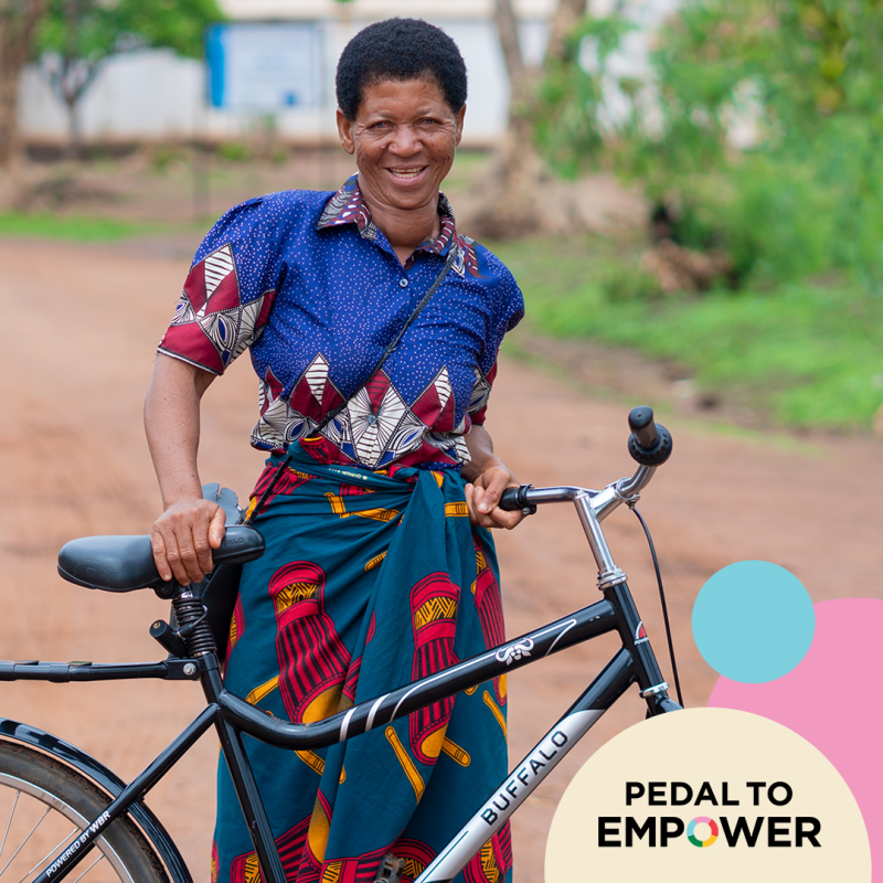 A smiling woman wearing a colorful shirt and a wrapped skirt stands next to a bicycle on a dirt path, promoting the Pedal To Empower campaign for World Bicycle Relief.