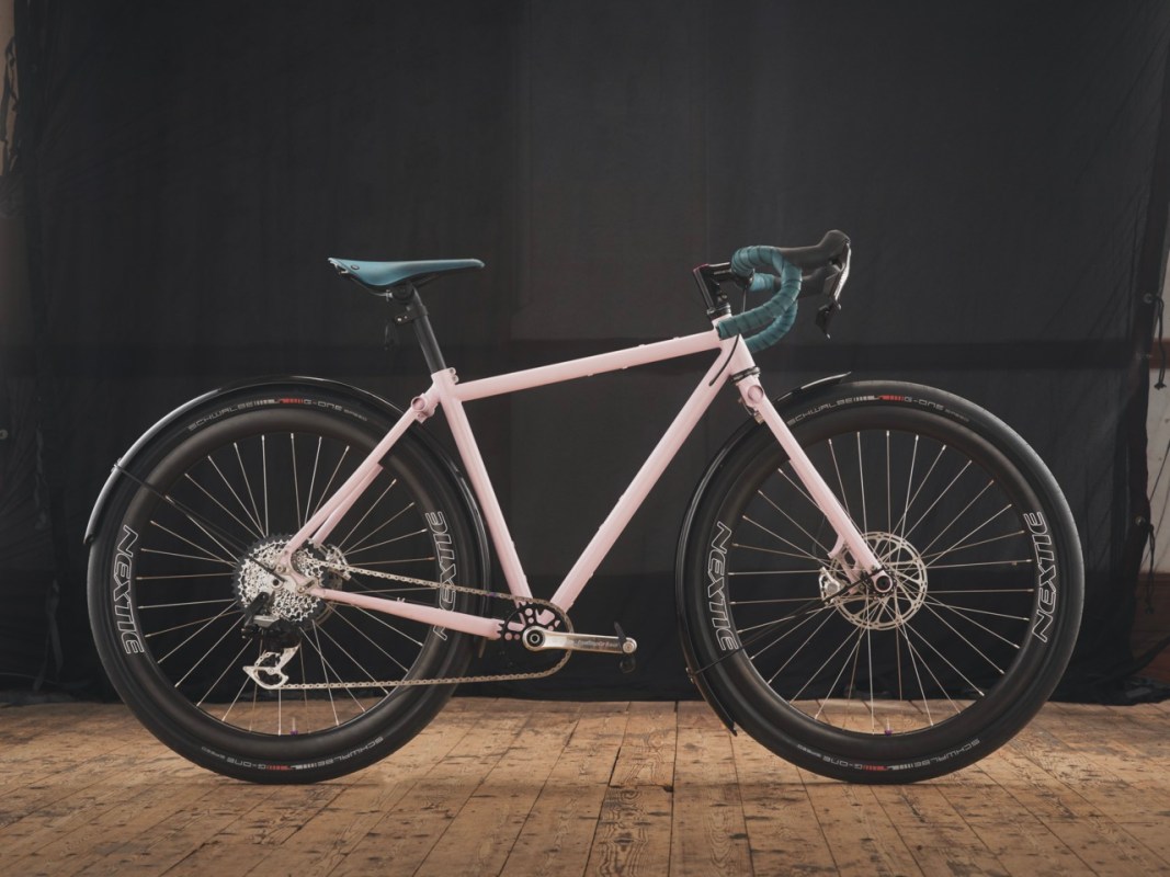 A pink bicycle with large black wheels and teal accents, positioned on a wooden floor against a dark background.