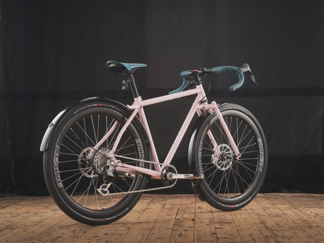 A pink bicycle with black wheels and fenders is displayed against a dark background, showcasing its modern design and features.