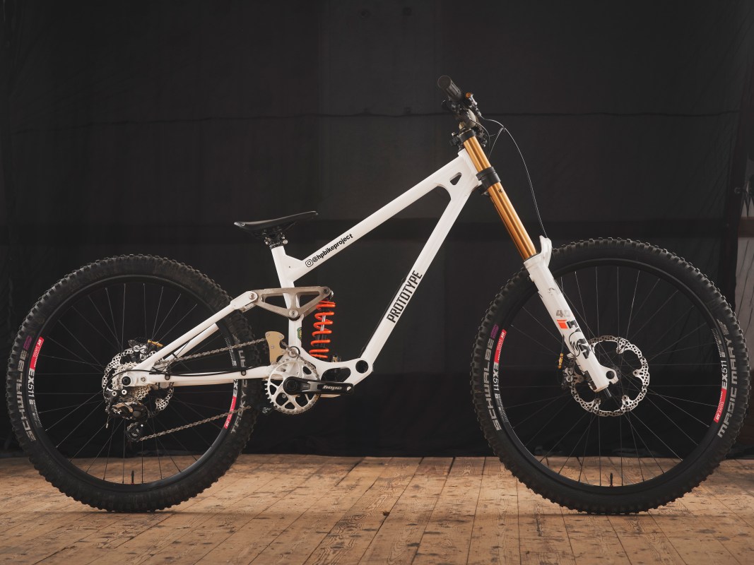 A custom downhill bike with a white steel frame, featuring orange suspension components and a unique design, showcased on a wooden floor against a black backdrop.