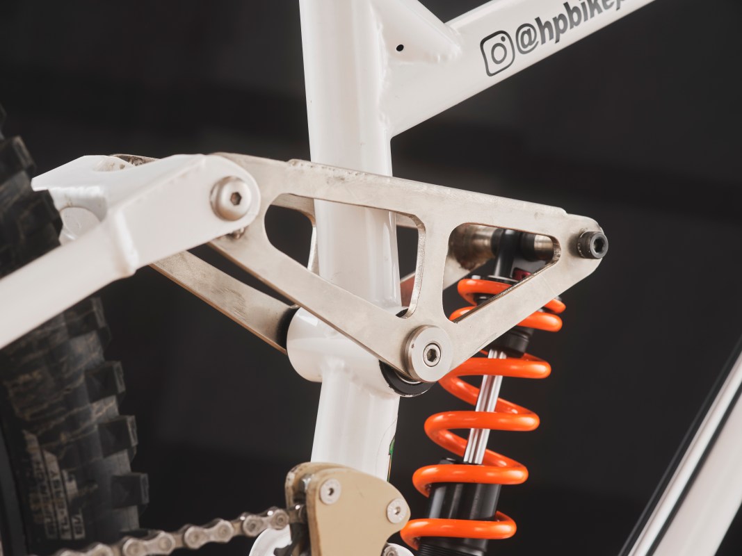 Close-up view of the rear suspension linkage and shock absorber on a custom downhill bike, featuring a steel frame and an orange coil spring.
