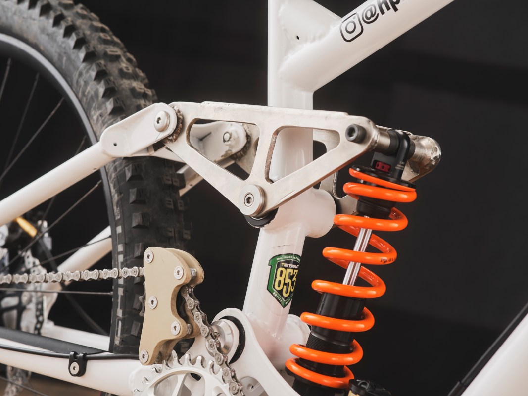 Close-up view of the rear suspension system of a custom downhill bike, featuring a white frame, orange shock absorber, and intricate linkage design.