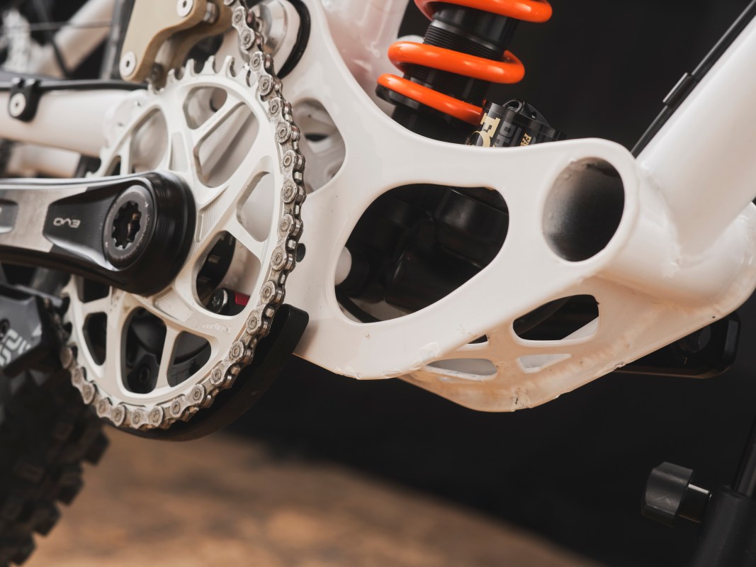 Close-up view of the drivetrain and suspension system of a custom downhill bike, featuring a white frame, crankset, chain, and orange shock absorber.