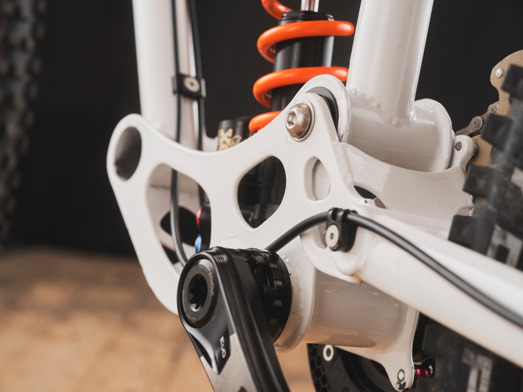 Close-up view of a downhill bike's bottom bracket section, featuring a white frame with a visible linkage system and an orange shock absorber.