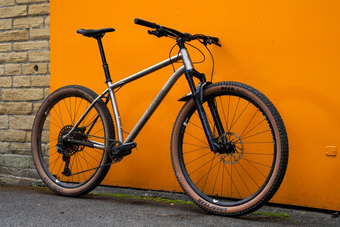 A Sonder Broken Road Ti mountain bike leaning against an orange wall, showcasing its sleek metallic frame and rugged tires, designed for bikepacking and long rides.