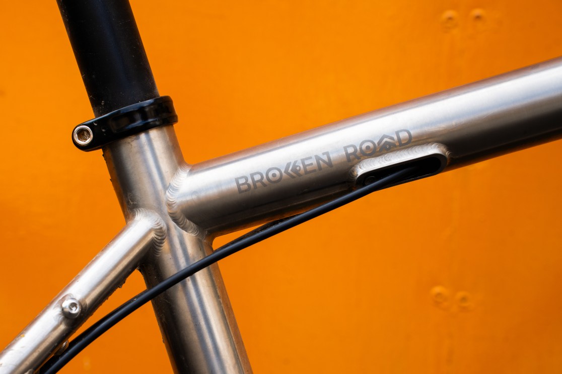 Close-up of the Sonder Broken Road Ti bike frame showcasing the etched logo and internal cable routing against an orange background.