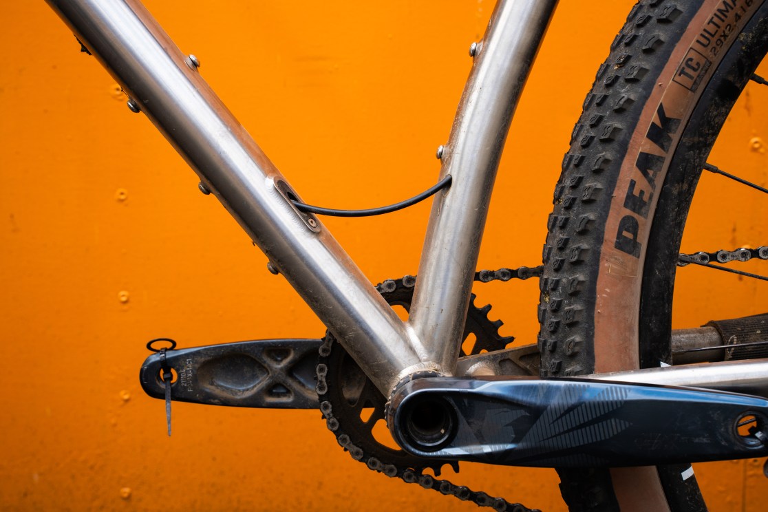 Close-up of the Sonder Broken Road Ti bike frame showing the bottom bracket, chainring, and internal cable routing against an orange background.
