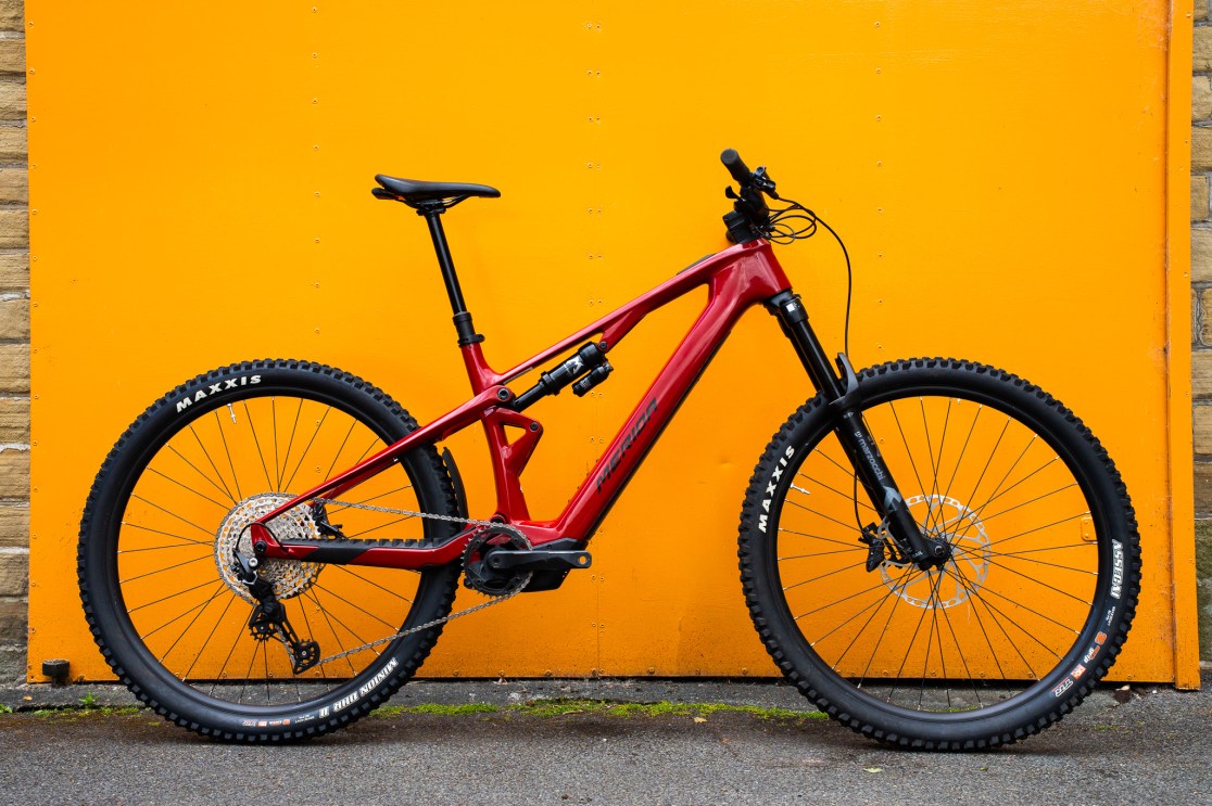Red mountain bike with dual suspension parked against an orange wall, featuring Maxxis tires and modern design.