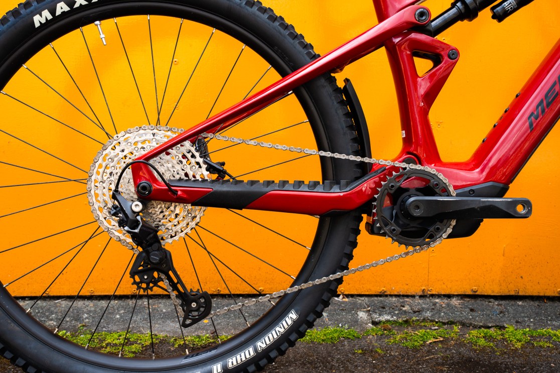 Close-up view of the rear drivetrain and chain of a Merida E160 mountain bike, featuring a red frame and a bright orange background.