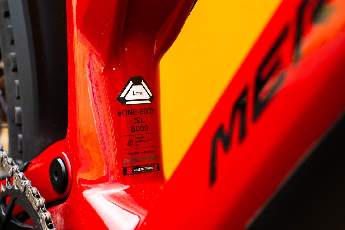 Close-up of the frame of a Merida eONE-SIXTY SL 6000 bike, featuring a bright red color with a label indicating it as a long frame. The text on the label includes designations like 'Designed in Germany' and 'Made in Taiwan.'