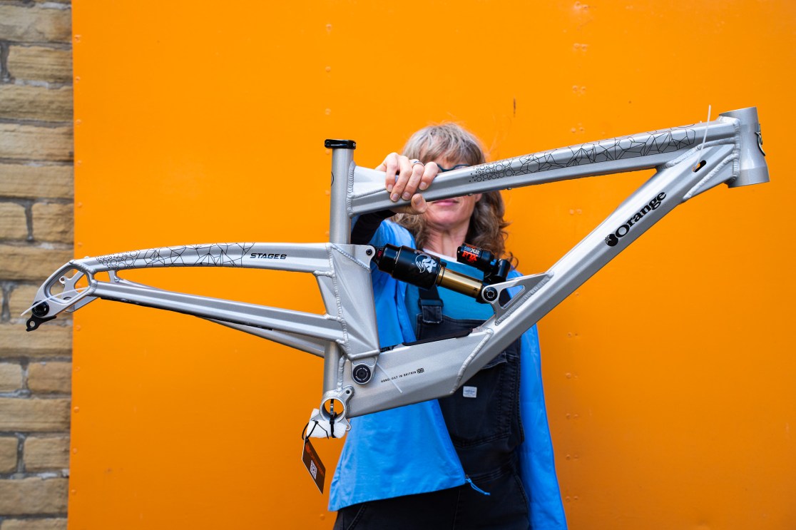 A person holding a silver mountain bike frame against an orange background.