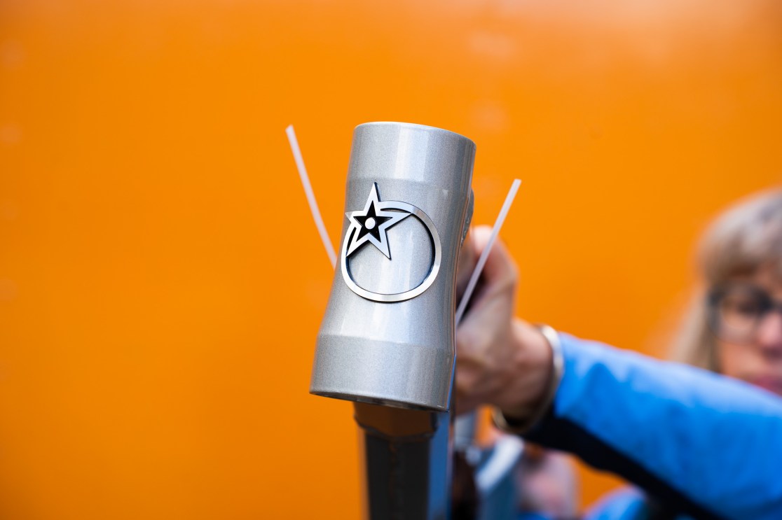 Close-up of a hand holding a metallic bicycle component with a star logo, against an orange background.
