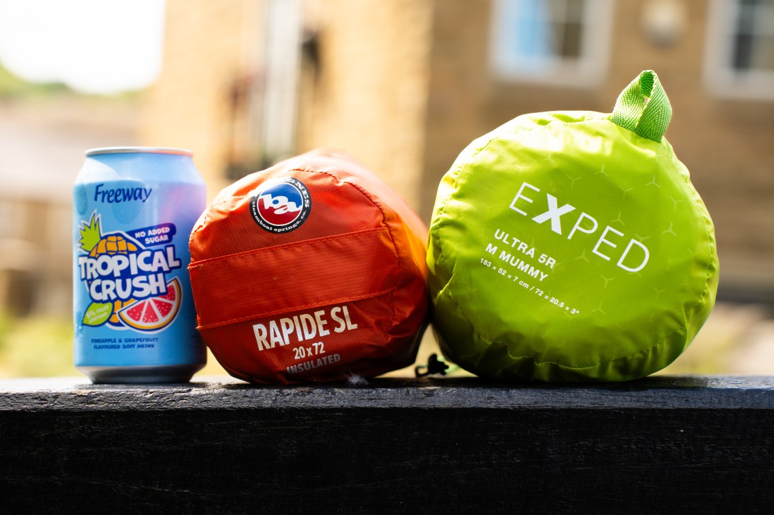 A can of Freeway Tropical Crush soda next to two packed sleeping mats: an orange Big Agnes Rapide SL and a green Exped Ultra 5R Mummy, resting on a wooden surface outdoors.