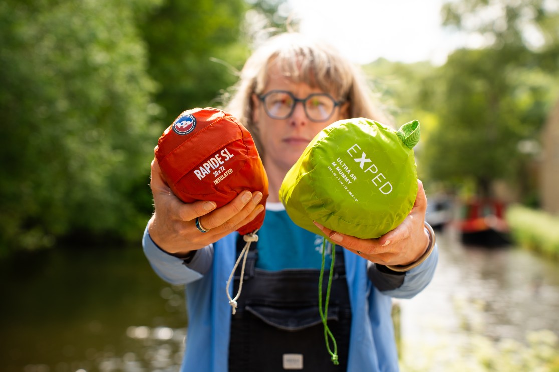 A person holds two sleeping mats, the Big Agnes Rapide SL (red) and the Exped Ultra 5R Mummy (green), against a blurred outdoor background by a canal.