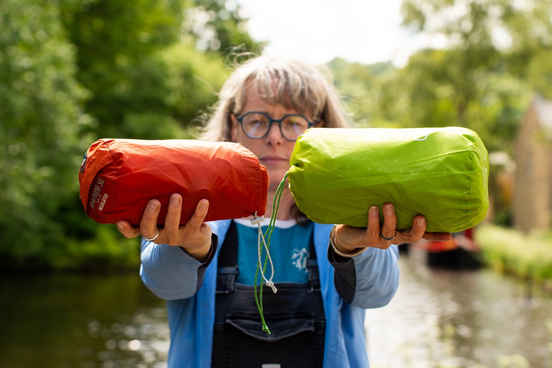 A person holding two insulated sleeping mats, one red and one green, while standing by a body of water surrounded by greenery.