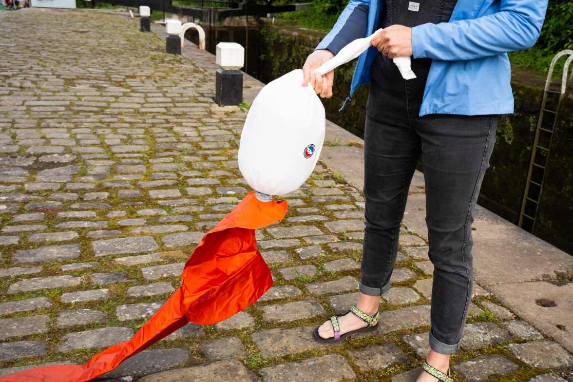 A person demonstrating how to use an inflatable dry bag, with a white and orange bag held in one hand while standing on cobbled ground next to a canal.
