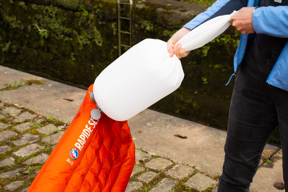 A person using an inflator bag to inflate a Big Agnes Rapide SL sleeping mat outdoors, with a cobblestone surface and greenery in the background.