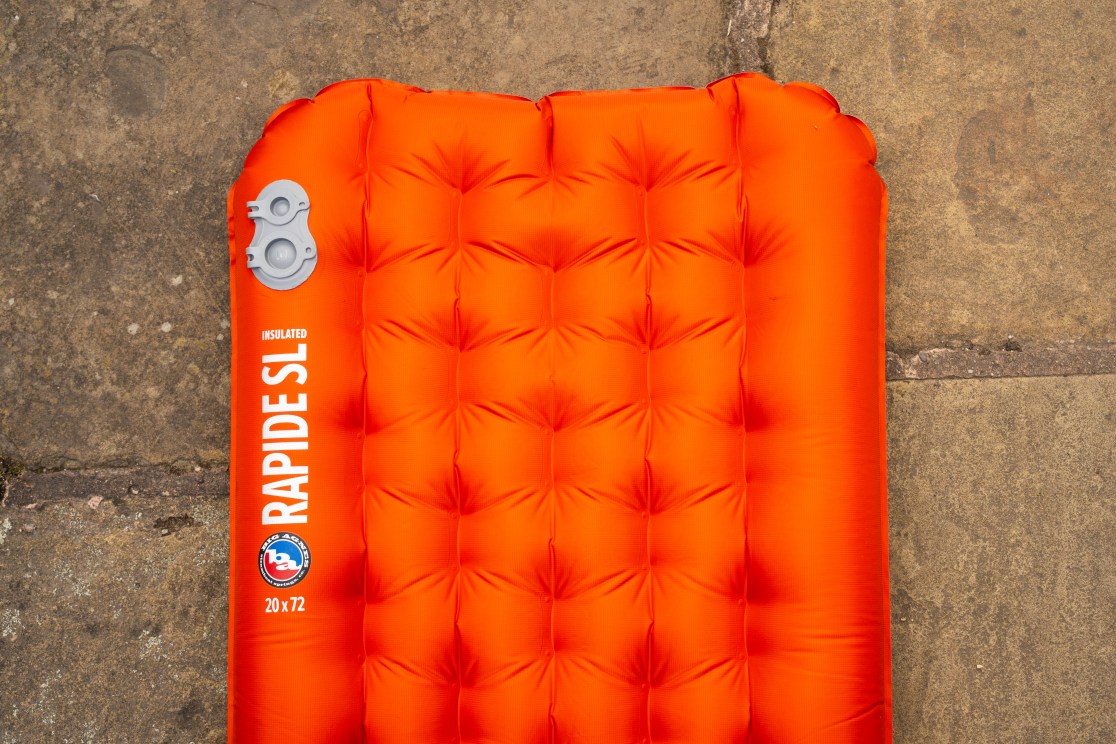 A close-up view of the Big Agnes Rapide SL insulated sleeping mat, featuring an orange color with buttons for inflation, showing its dimensions 20x72 inches.