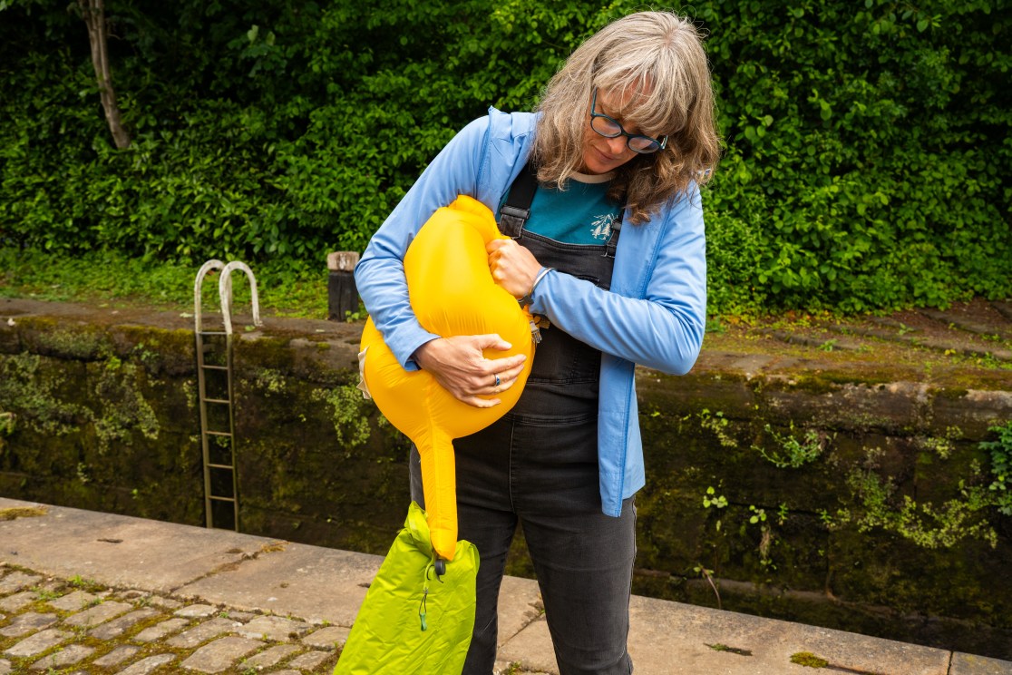 A person inflating a yellow sleeping mat while holding it under their arm, with greenery in the background.