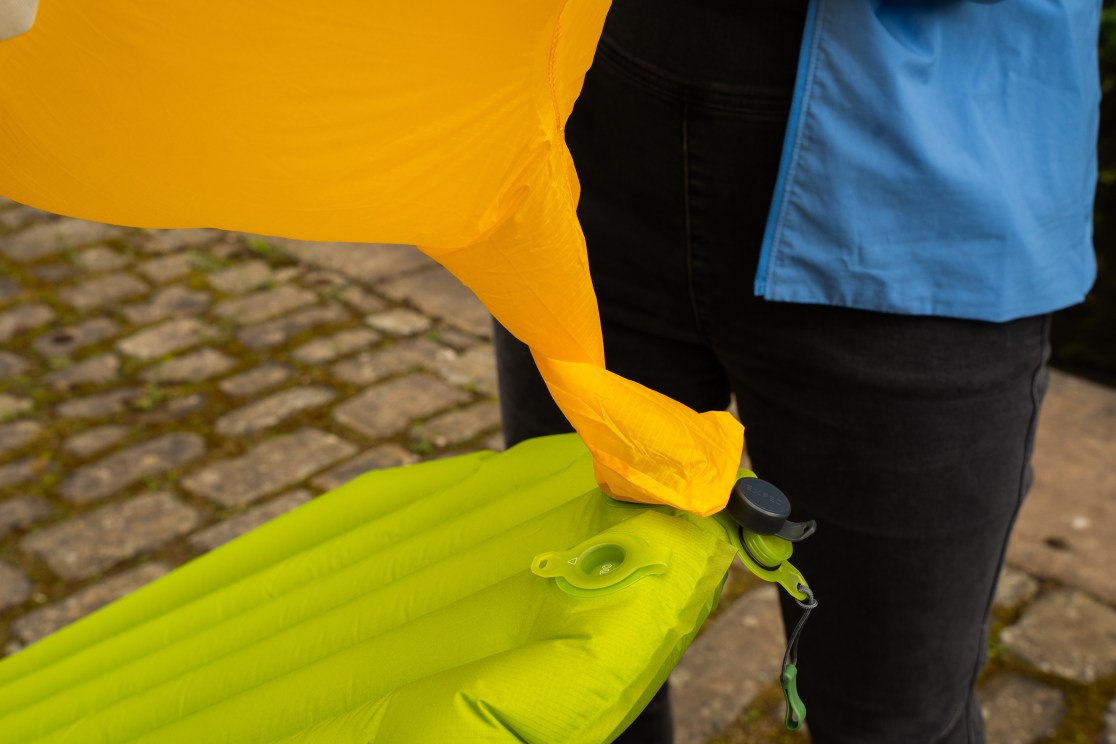 A close-up of a person inflating a green sleeping mat using an orange inflator bag, set on a cobblestone surface.