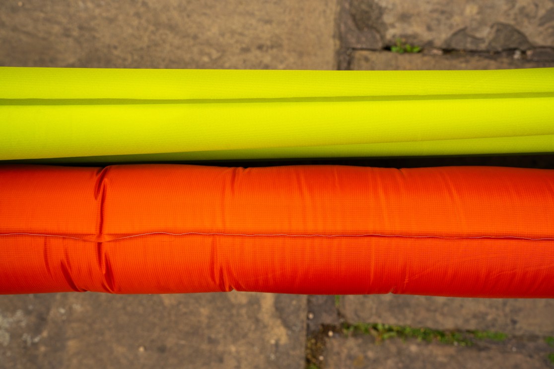 Two inflatable sleeping mats placed side by side; one is bright green and the other is orange, resting on a textured stone surface.