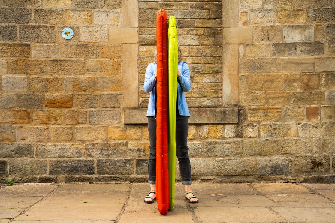 A person holding two insulated camping mats, one red and one green, in front of a stone wall.