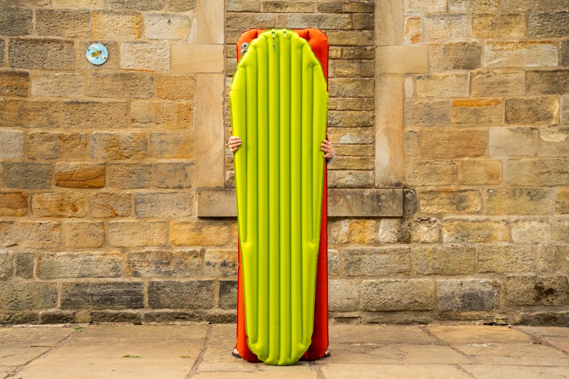 A person holding a large, inflatable mat with a bright green top and orange bottom, standing against a textured stone wall.
