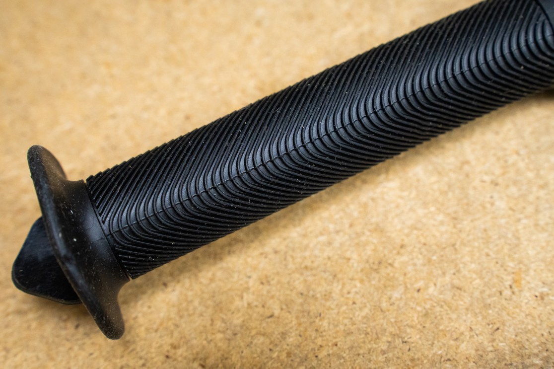 Close-up of a black bike grip with textured surface and flange on one side, placed on a wooden background.
