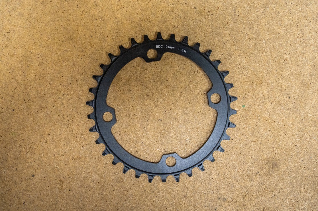 Black chainring with a 104mm BDC and a 34-tooth design, displayed on a wooden surface.