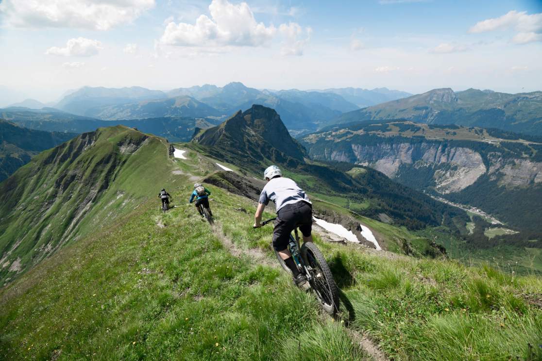 Three mountain bikers riding on a grassy mountain trail overlooking scenic valleys and peaks under a partly cloudy sky.