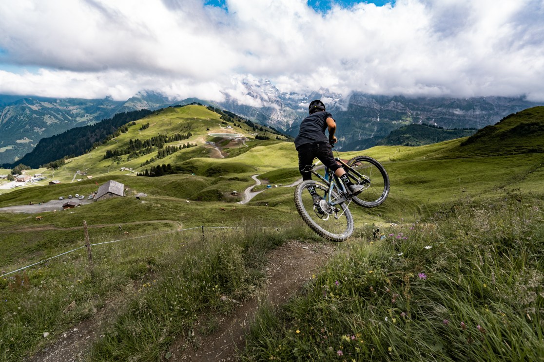 A mountain biker performing a wheelie on a trail surrounded by green hills and distant mountains under a cloudy sky.