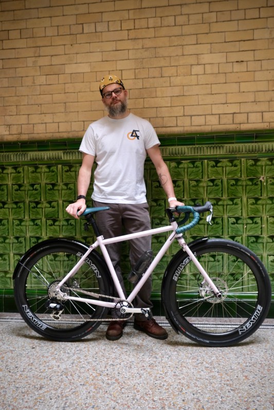 A bearded man wearing a T-shirt and cap stands next to a unique bike in front of a decorative green tiled wall.