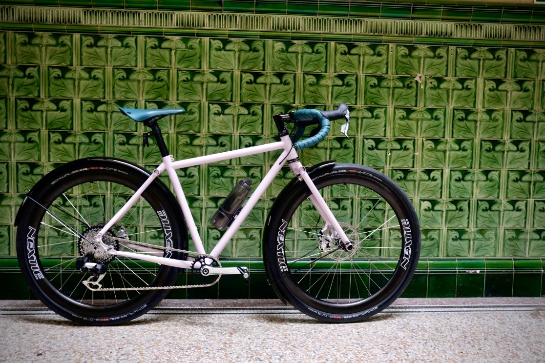 Side view of a unique froad bike with fat tires, showcasing a light pink frame, green handlebar grips, and a sleek design, set against a decorative green tiled wall.