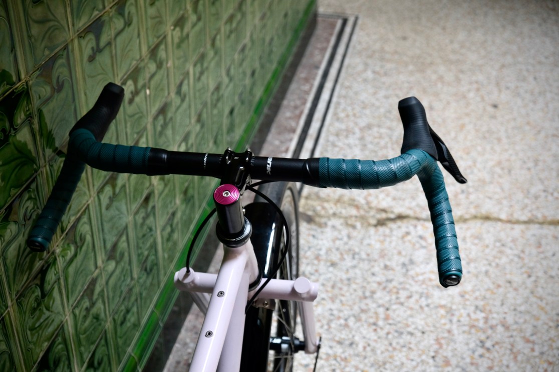 Close-up view of the handlebars of a custom bike with teal grips, a pink headset cap, and a sleek design against a patterned green wall.