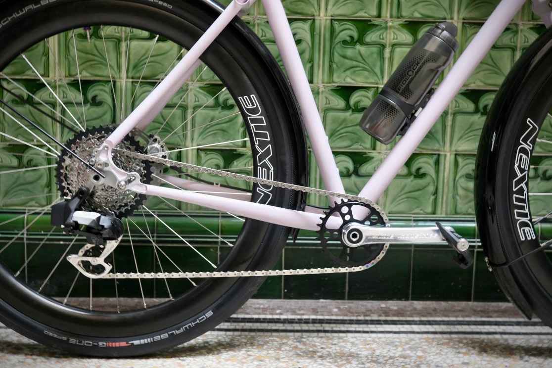 Close-up of a pink gravel bike showcasing its drivetrain and fat tires, positioned against a patterned green tile background.