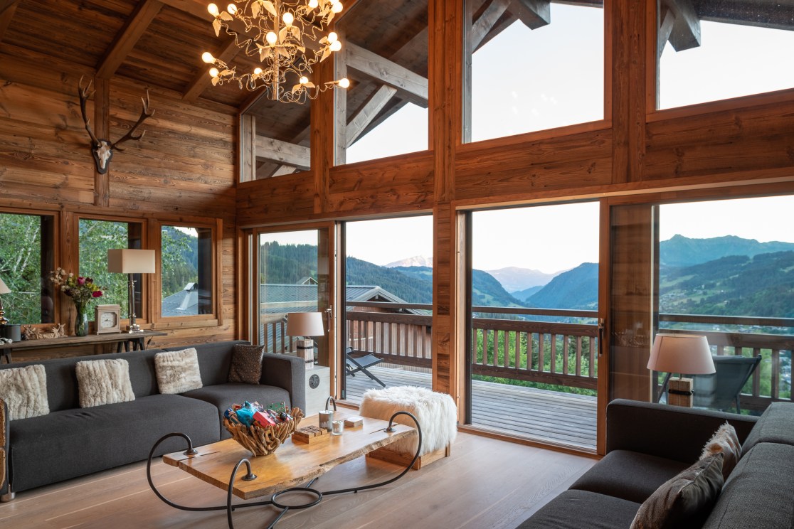 Interior of a cozy mountain chalet featuring wooden walls, a comfortable gray sofa with decorative cushions, a rustic wooden coffee table, and large windows showcasing a view of mountains and greenery outside.