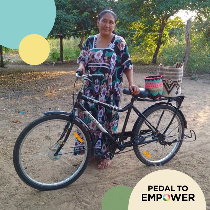 A woman stands beside a bicycle in a natural outdoor setting, holding a basket. She is wearing a floral-patterned dress, and there are colorful circular designs in the corners of the image, emphasizing the theme of empowerment.