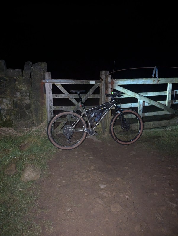 A Sonder Broken Road Ti mountain bike leaning against a wooden gate at night, with gravel path and grassy surroundings.
