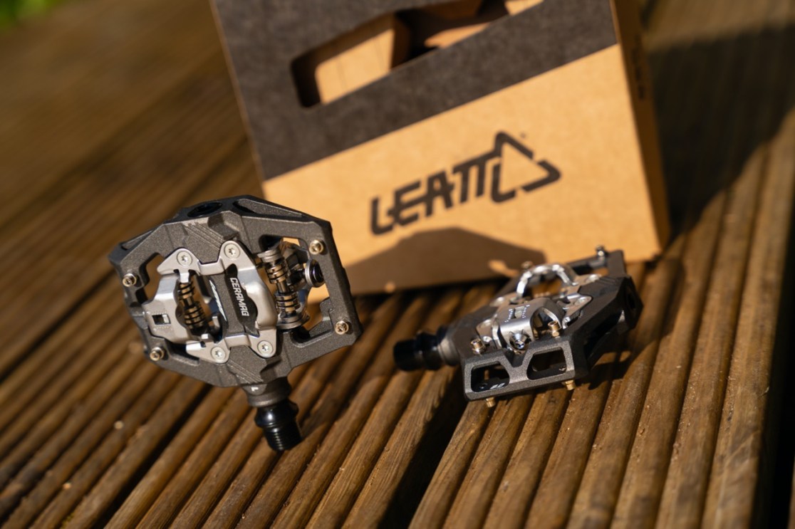 Two bicycle pedals displayed on a wooden surface, with a cardboard box labeled 'Leatt' in the background.