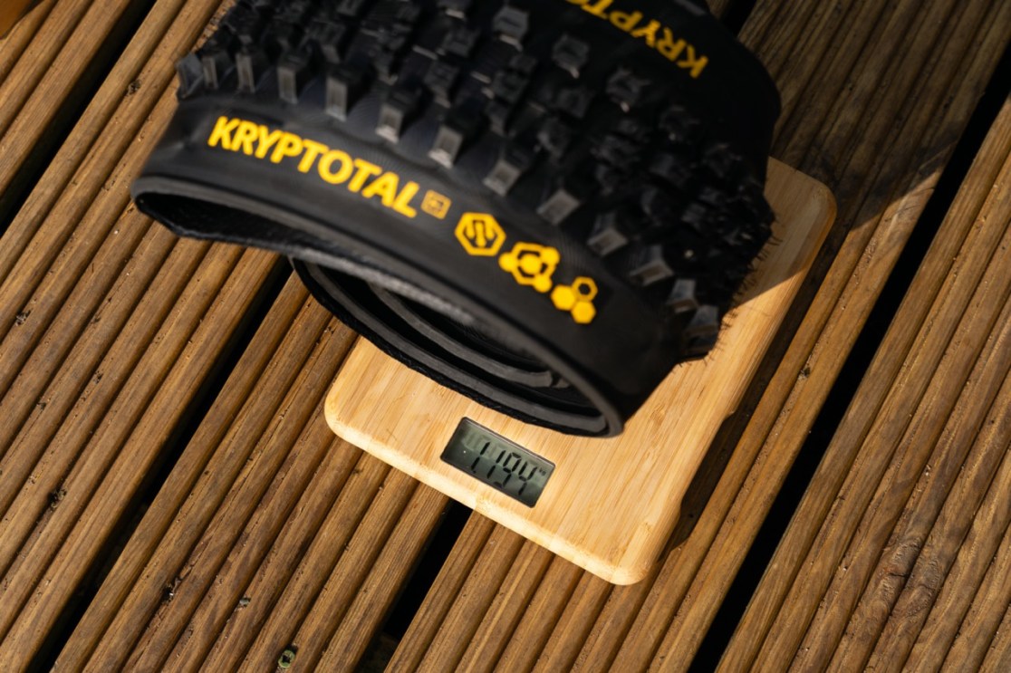 A close-up image of a KRYPTOTAL bicycle tire placed on a digital scale, showing a weight of 1194 grams. The background features wooden planks.